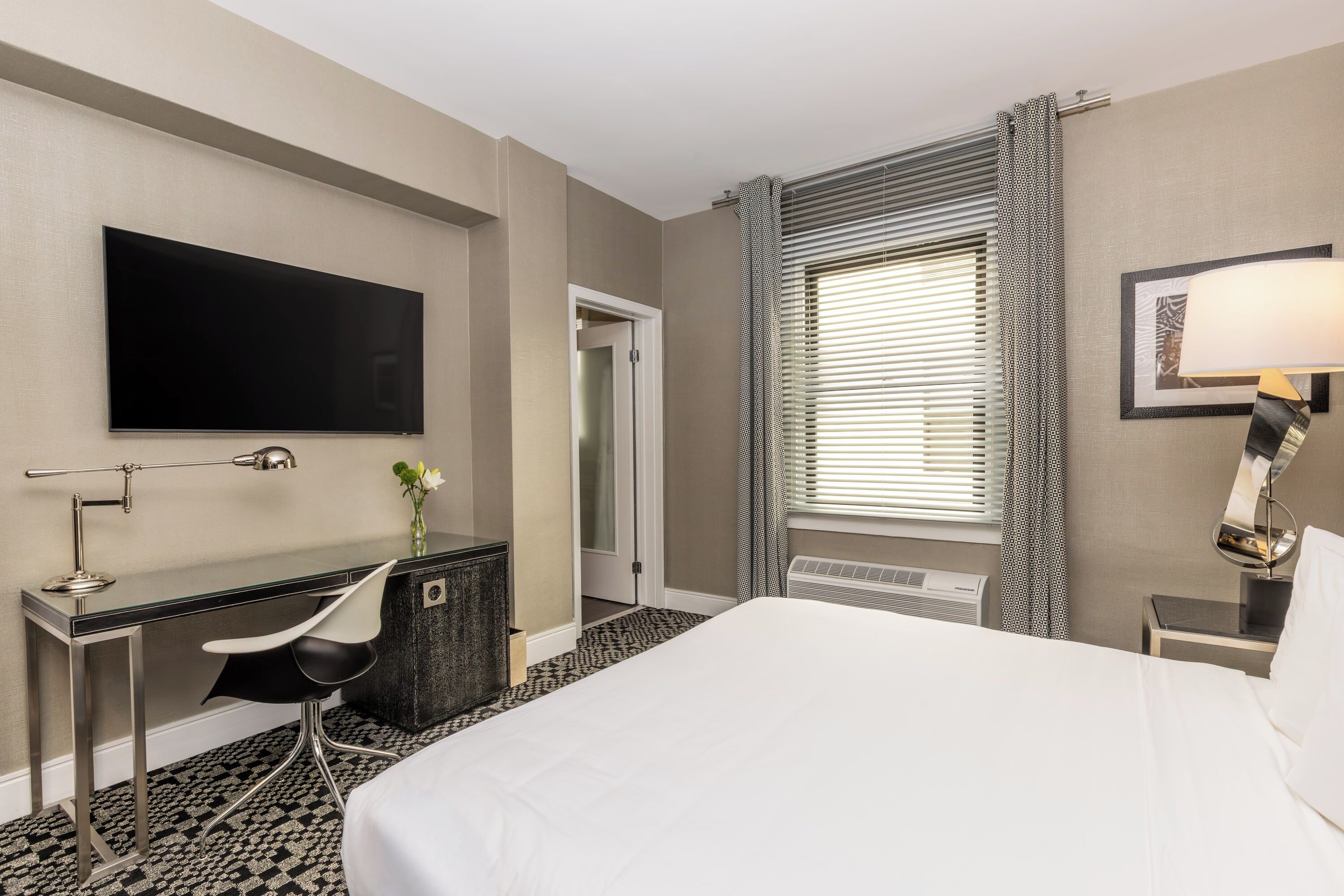 A hotel room with a bed, desk, chair, TV, and a window with blinds.