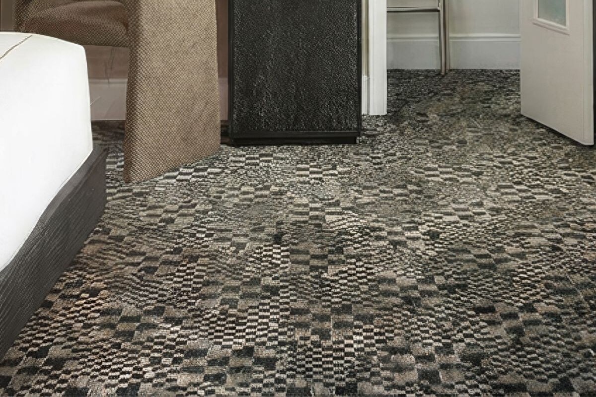 A hotel room with a bed, a desk with a lamp, a chair, a small refrigerator, a wardrobe, and a checkered carpet.
