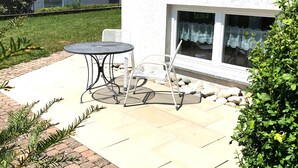 Une terrasse avec une table ronde et deux chaises, un petit jardin avec des plantes et un bâtiment doté d’une fenêtre.