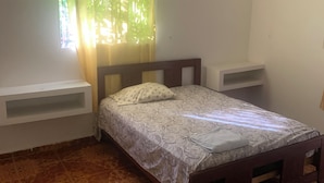 Ein Schlafzimmer mit einem Bett, einem Fenster mit Vorhängen, einem weißen Regal und fußbodenhohen, gemusterten Fliesen.