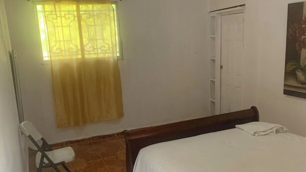 Un dormitorio con cama, una silla, un ventanal con cortina y una puerta.
