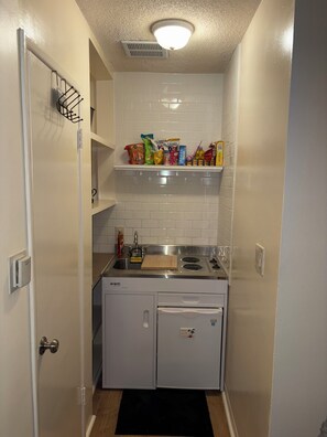 A compact kitchen with a dishwasher, a stove, and a small refrigerator. There are shelves above the sink holding various food items. - Modern & Private Studio Retreat in the Heart of Inglewood (Inglewood)