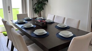 Un coin repas avec une table en bois foncé, dressée pour six personnes, entourée de chaises blanches. Un pot de fleurs se trouve dans un coin, et un panier contenant divers objets est posé sur la table.