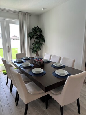 A dining area with a dark wooden table set for six, surrounded by white chairs. A potted plant is in the corner, and a basket with items is on the table. - Disney World 15min! Private Pool 4 Bedroom Themed House! (Davenport)