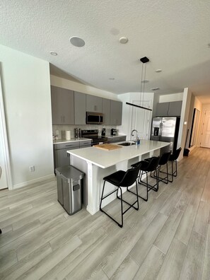 A modern kitchen with grey cabinets, a stainless steel refrigerator, a microwave, and a large island with bar stools. - Disney World 15min! Private Pool 4 Bedroom Themed House! (Davenport)