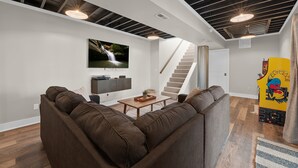 Una sala de estar moderna con un sofá marrón, una mesa de centro y un televisor montado en la pared. Se ve una escalera que sube a otro nivel y una obra de arte colorida en la pared.