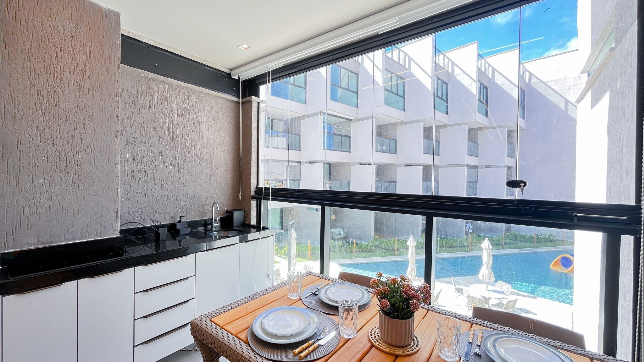A modern kitchen with a large window, a dining area with a table set for four, and a view of a swimming pool and buildings.