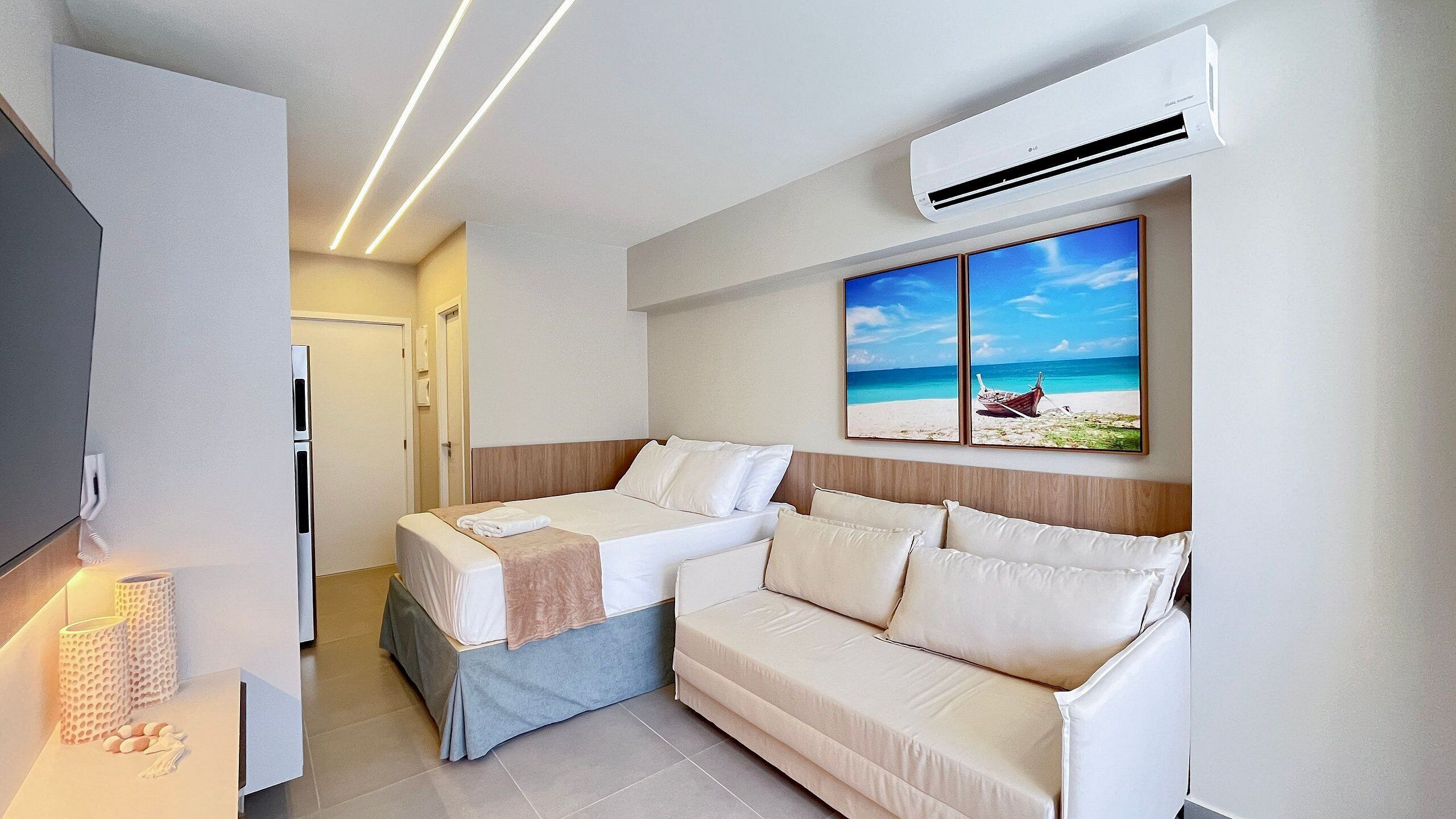 A hotel room with a bed, a sofa, an air conditioner, a painting of a beach, and a phone on the wall.