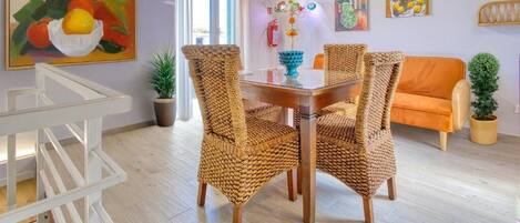A dining area with wicker chairs, a wooden table, and a painting of fruit on the wall.