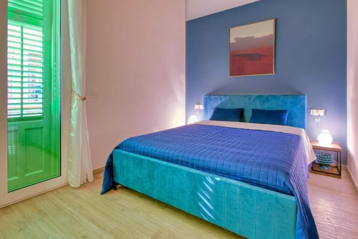 A bedroom with a bed, a nightstand, a wall-mounted lamp, and a window with blinds.