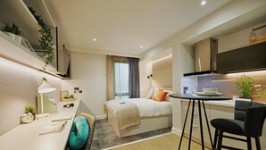 A compact hotel room with a bed, a desk, a chair, a small kitchenette, and a window with curtains. - Student Only West End (Glasgow)