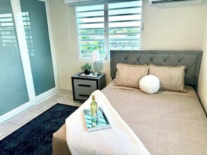 A bedroom with a bed, a bedside table with a lamp, and a window with blinds. - Luxe Black & White Apt I (Trujillo Bajo)