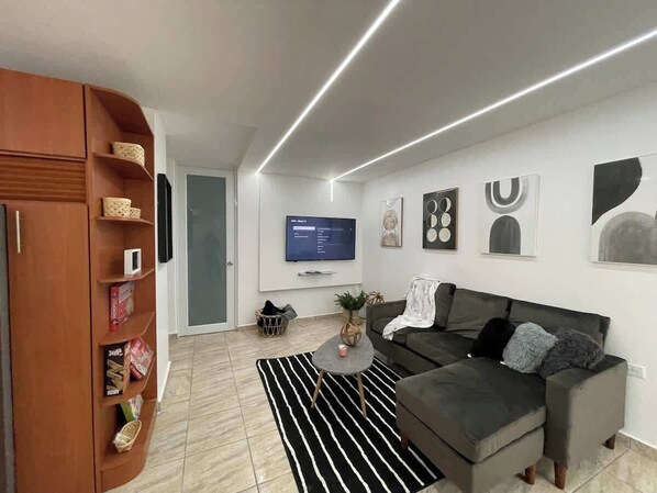 Un salon moderne avec un canapé d’angle gris, un tapis rayé noir et blanc, un meuble en bois avec rangements, un téléviseur à écran plat fixé au mur et une petite table basse ronde.