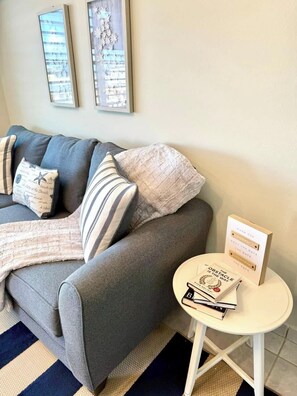 A grey sofa with striped and starfish-patterned cushions, a small white side table with a book and a motivational card, and a blue and beige striped rug. - Luxe Black & White Apt II (Carolina)