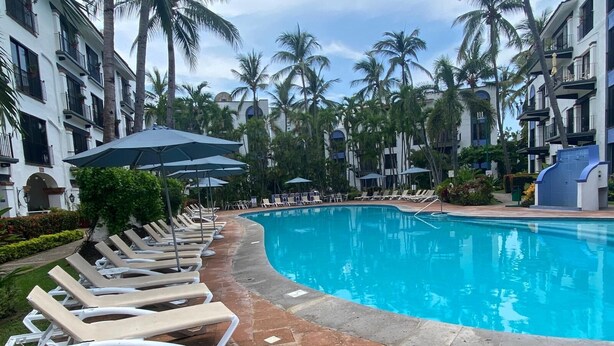 Une piscine entourée de chaises longues et de parasols, bordée de palmiers et d’un bâtiment doté de balcons.