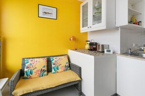 A compact kitchen with white cabinets, a small dining area with a wicker chair and a yellow cushion, and a framed picture on the yellow wall. - Ajoncs Apartment – Seafront Terrace, Parking, Côte Sauvage (Batz-sur-Mer)