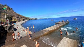 Ein Strand mit badenden und sonnenden Personen, einer felsigen Küste und einem klaren, blauen Himmel.
