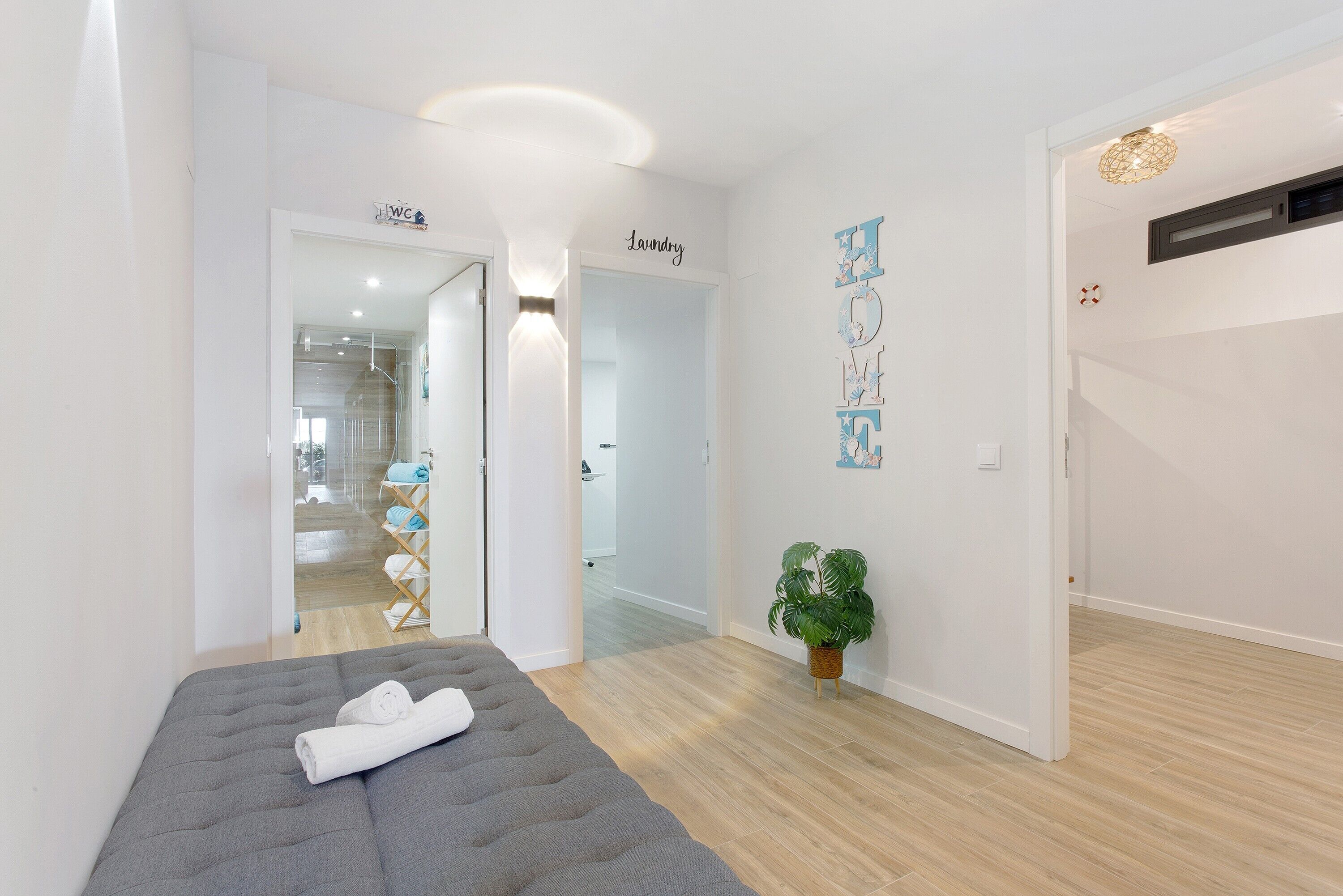 Une chambre d’hôtel moderne avec un lit gris, une plante en pot et un couloir menant à la salle de bain et à l’espace buanderie.