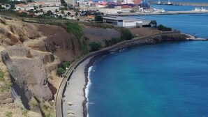 Eine Küstenstraße mit einem Strand rechts, einem felsigen Klippenhang links und Gebäuden im Hintergrund.