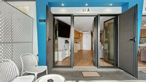 Ein moderner Hauseingang zu einer Wohnung, durch Glastüren ist eine Küche sichtbar, sowie ein Balkon mit Sitzgelegenheit und eine Nummer am Türschild.