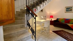 Una escalera con barandilla de metal conduce a un rellano. En el rellano hay un escritorio blanco con una lámpara y una silla. Un sofá de colores se ve al fondo.