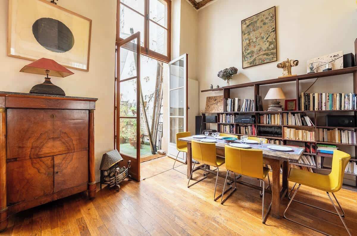 A room with a wooden cabinet, a bookshelf, a dining table with yellow chairs, and a large window.