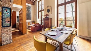 Een eethoek met een houten tafel gedekt voor vier personen, omringd door gele stoelen, in een kamer met zichtbare houten balken en grote ramen.