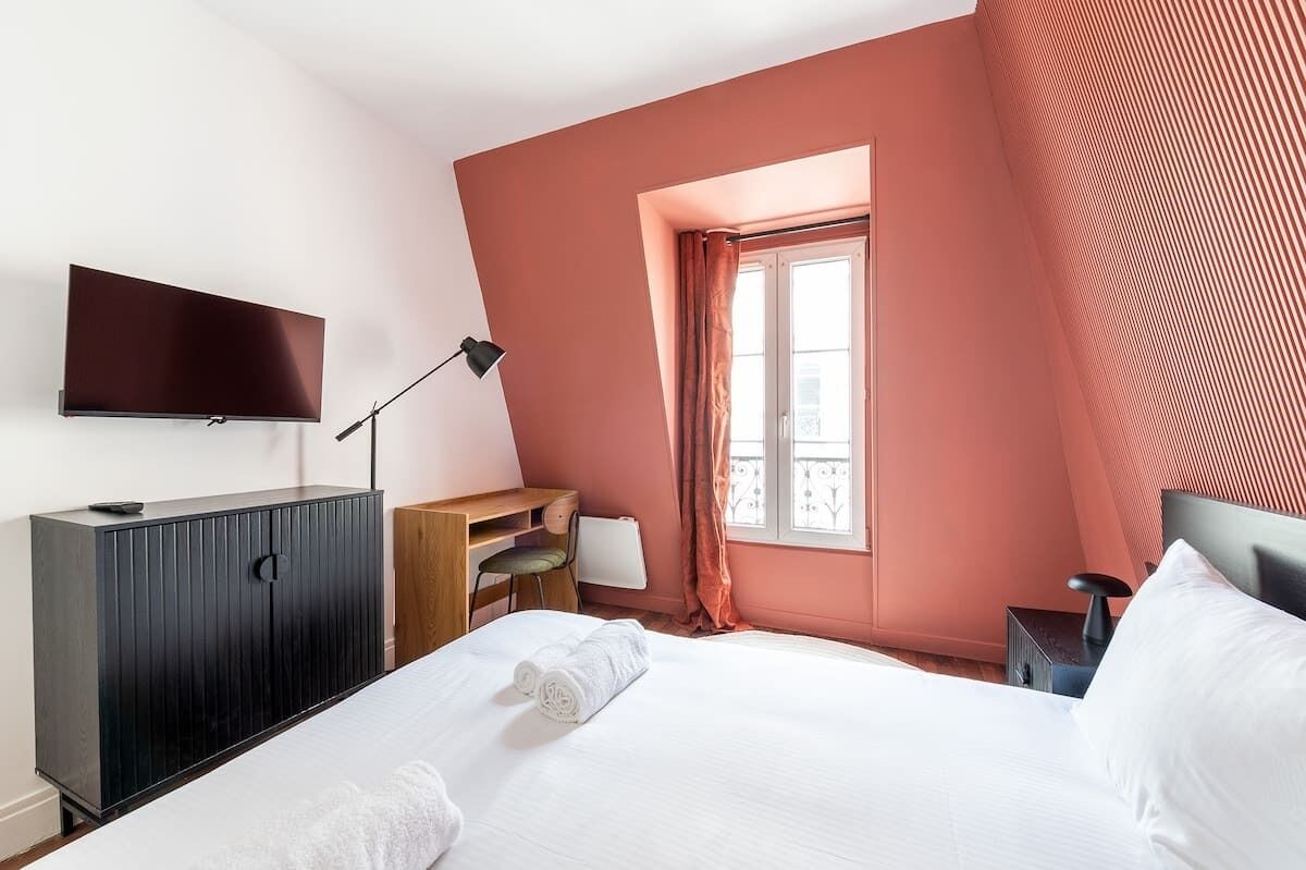 A bedroom with a bed, a TV, a desk, a chair, and a window with curtains.