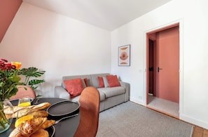 Ein Wohnzimmer mit einer Couch, einem Esstisch mit Brot und einer Blumenvase, und einer Tür rechts.