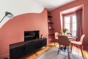 Ein Wohnzimmer mit Fernseher auf einer schwarzen Kommode, einem Esstisch mit Stühlen, einem Fenster mit Vorhängen und einer kleinen Pflanze auf der Kommode.