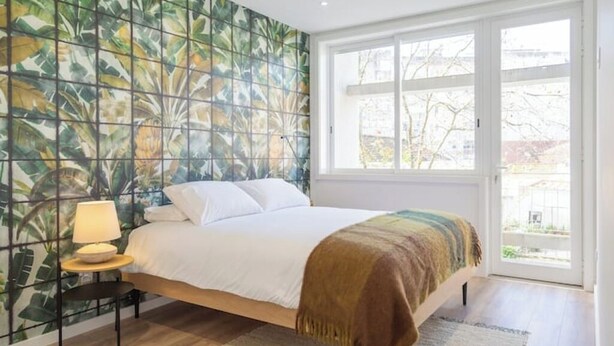 Un dormitorio con cama, mesita de noche, lámpara, ventana y vistas al exterior.