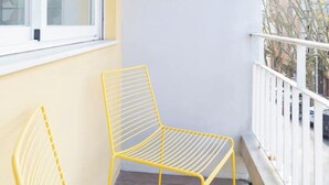 Una silla de metal amarillo en un balcón con barandilla blanca, una ventana y vistas a edificios y árboles.