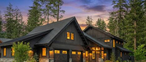 Une grande maison en bois sombre, dotée de plusieurs portes de garage et d’un toit à forte pente, entourée d’une pelouse bien entretenue et de grands arbres.