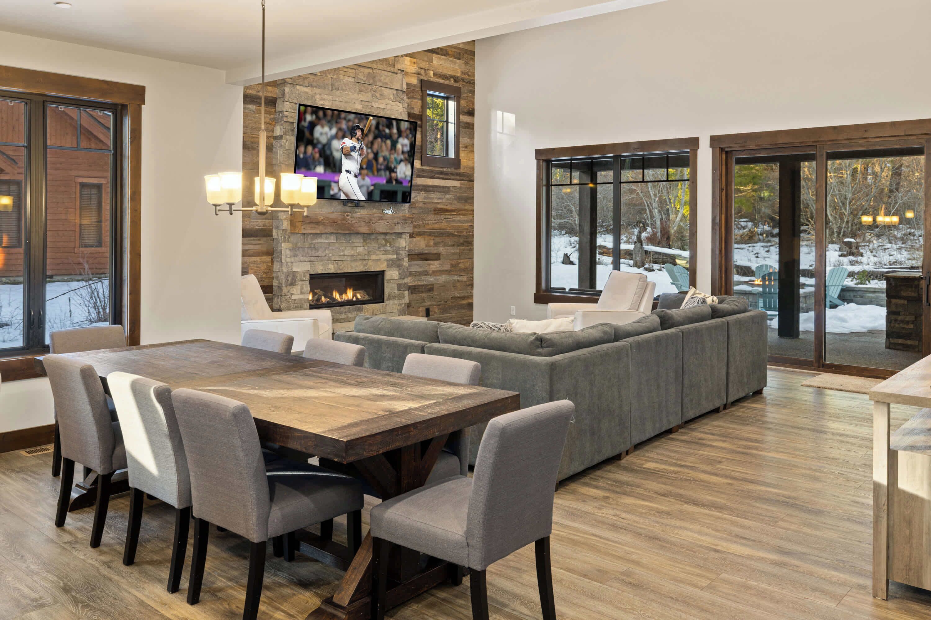 Ein modernes Wohnzimmer mit Kamin aus Stein, einem großen, darüber angebrachten Fernseher, einem grauen Ecksofa, einem Holzessttisch mit Stühlen und großen Fenstern mit Blick ins Freie.