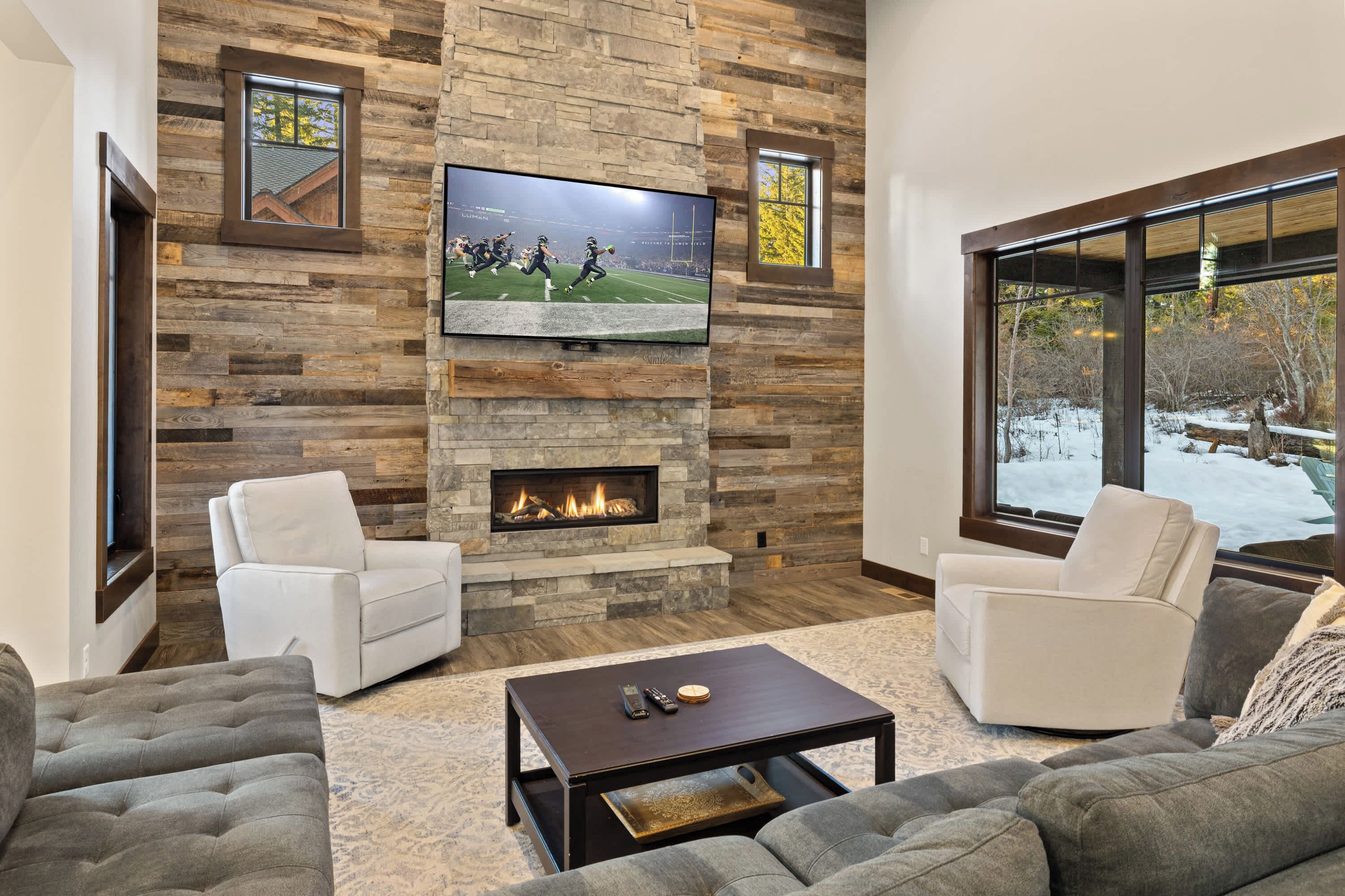 A modern living room with a stone fireplace, a flat-screen TV, and a large window with a view of a snowy landscape.