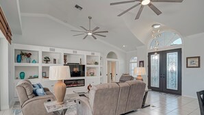 Un ampio soggiorno con un divano angolare beige, un tavolino e una televisione. Si vedono scaffali bianchi con oggetti decorativi e una ventola a soffitto.