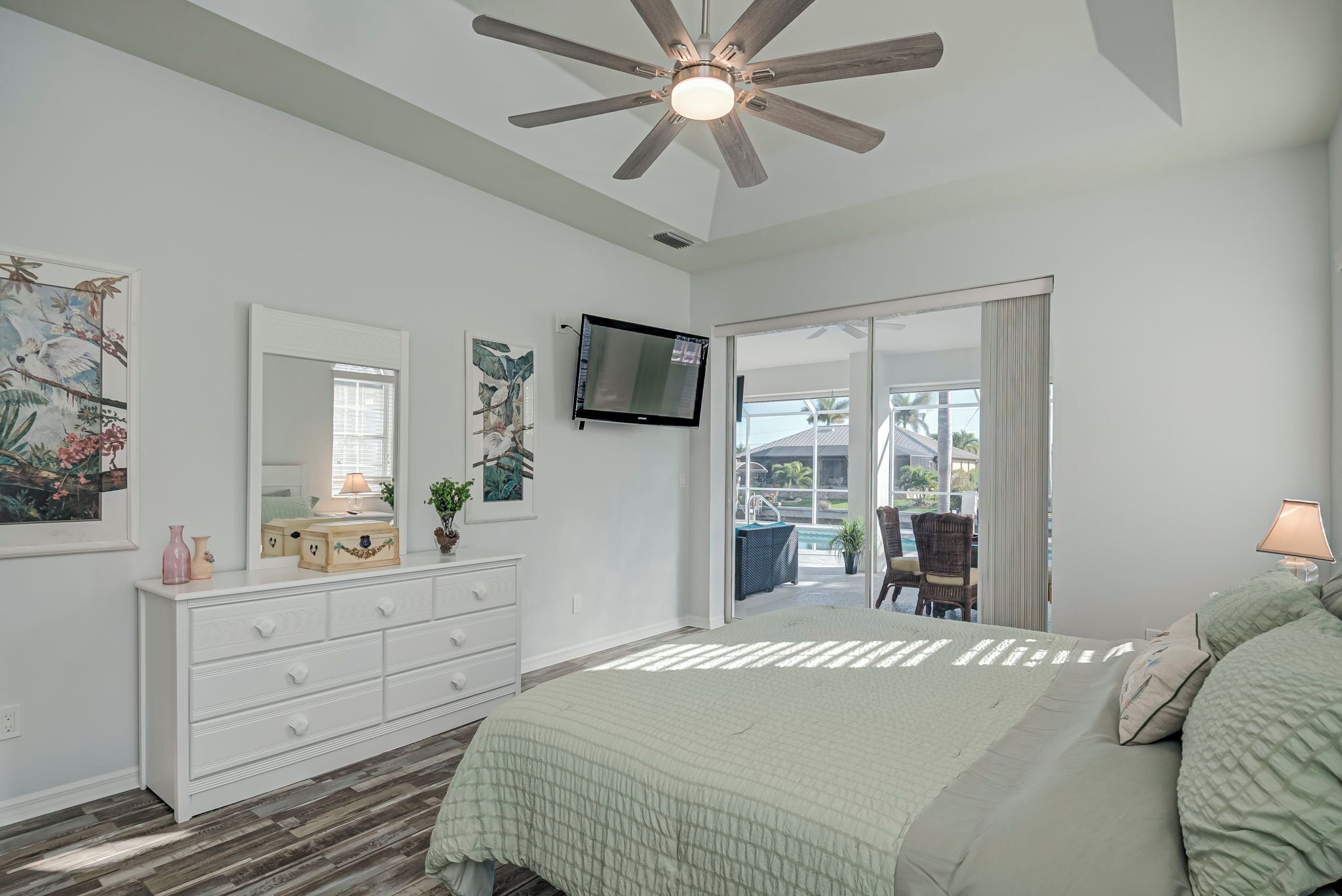 Un dormitorio con cama, ventilador de techo, televisor, cómoda y una ventana grande con vistas al exterior.