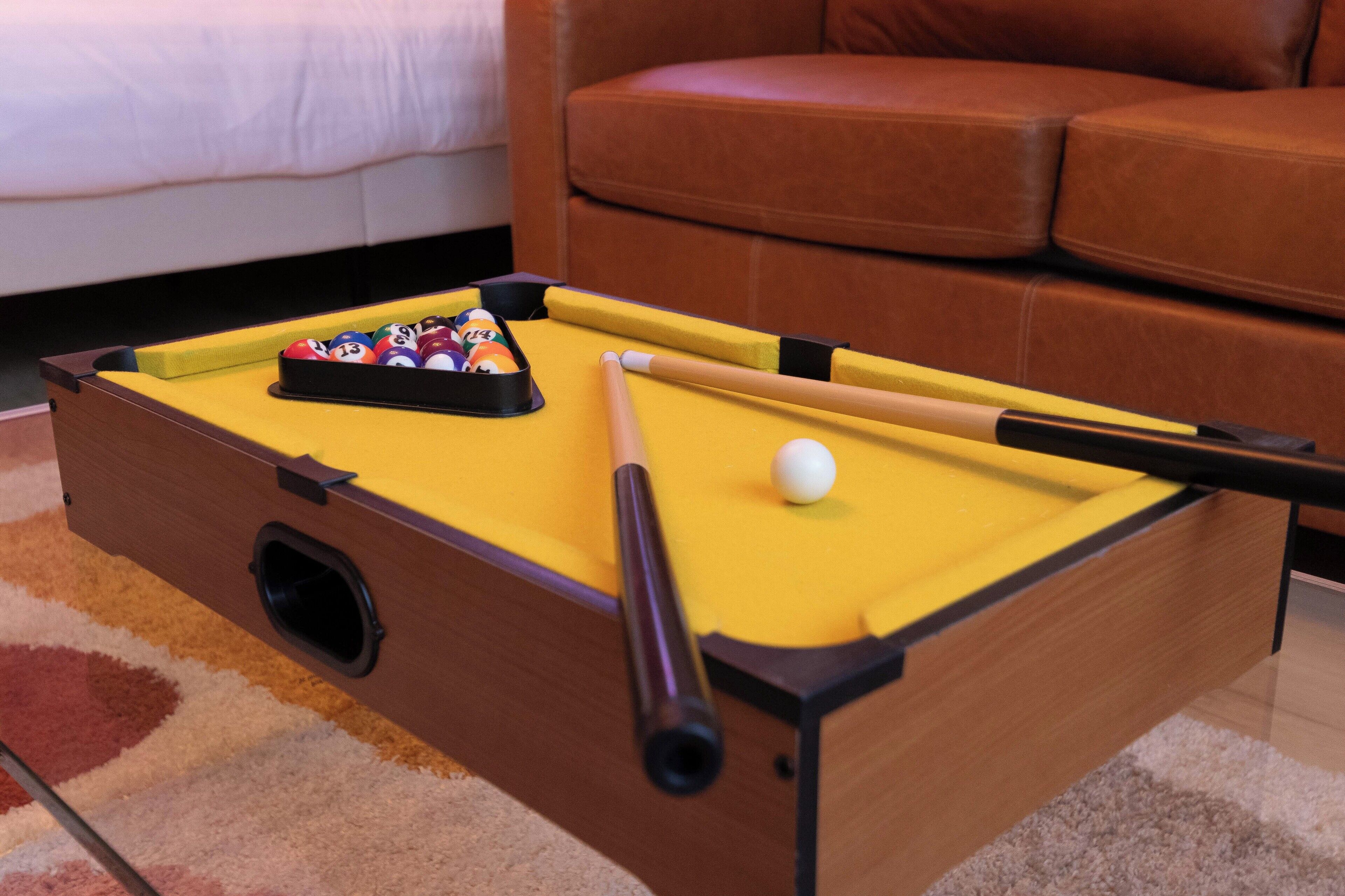 A pool table with a yellow felt surface, a set of billiard balls, and a cue stick.