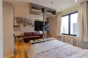 A modern hotel room with a bed, a desk, a TV, a sofa, and a window with a view of the city. - Moderno Studio nos Jardins Funcional e com Lazer (São Paulo)