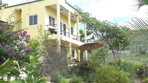A two-story yellow building with balconies, surrounded by lush greenery and flowering plants. - Escape Casita – Incredible Ocean Views, A/C & Heated Saltwater Pool (Todos Santos)