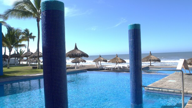En pool med kristallklart blått vatten, omgiven av solstolar och parasoller med halmtak, med utsikt över havet.