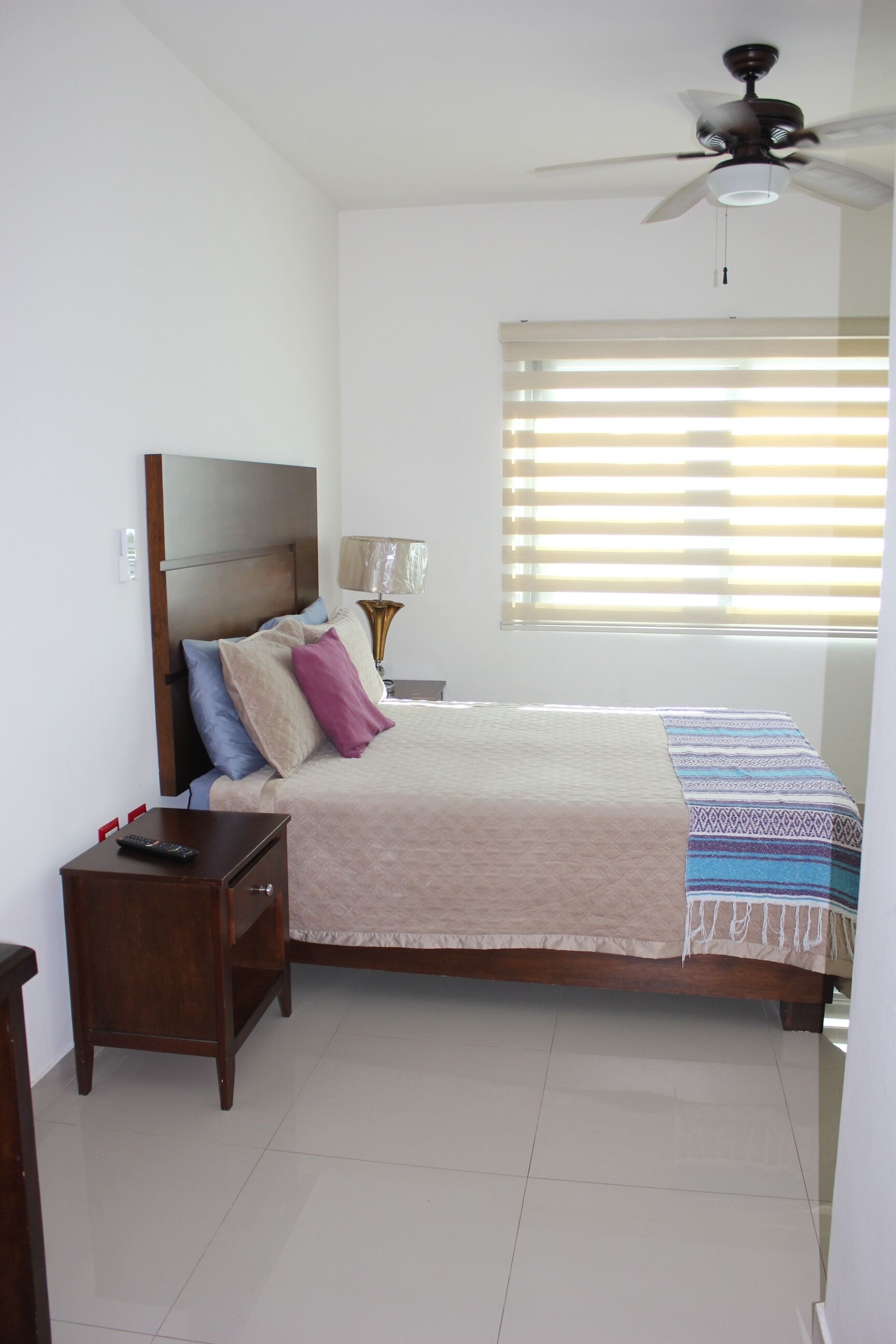 A bedroom with a bed, bedside table, lamp, ceiling fan, and window with blinds.