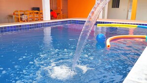 En swimmingpool med en vattenfontän, omgiven av en byggnad med orange väggar och en terrass med kakelplattor.
