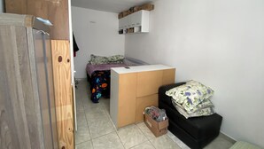 Une petite pièce, de taille modeste, comprenant un lit, un bureau et un canapé noir. Un placard en bois se trouve à gauche, et une petite étagère avec des boîtes est fixée au mur.