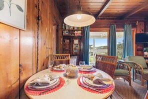 Interior de madera con una mesa de comedor dispuesta para cuatro personas, rodeada de sillas de mimbre, una estantería y un televisor.