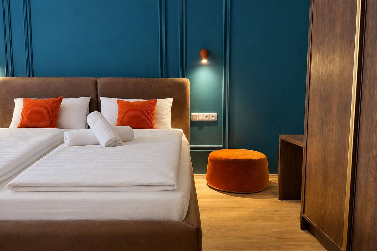 A double bed with white linens and orange pillows, a wooden floor, and a teal accent wall.