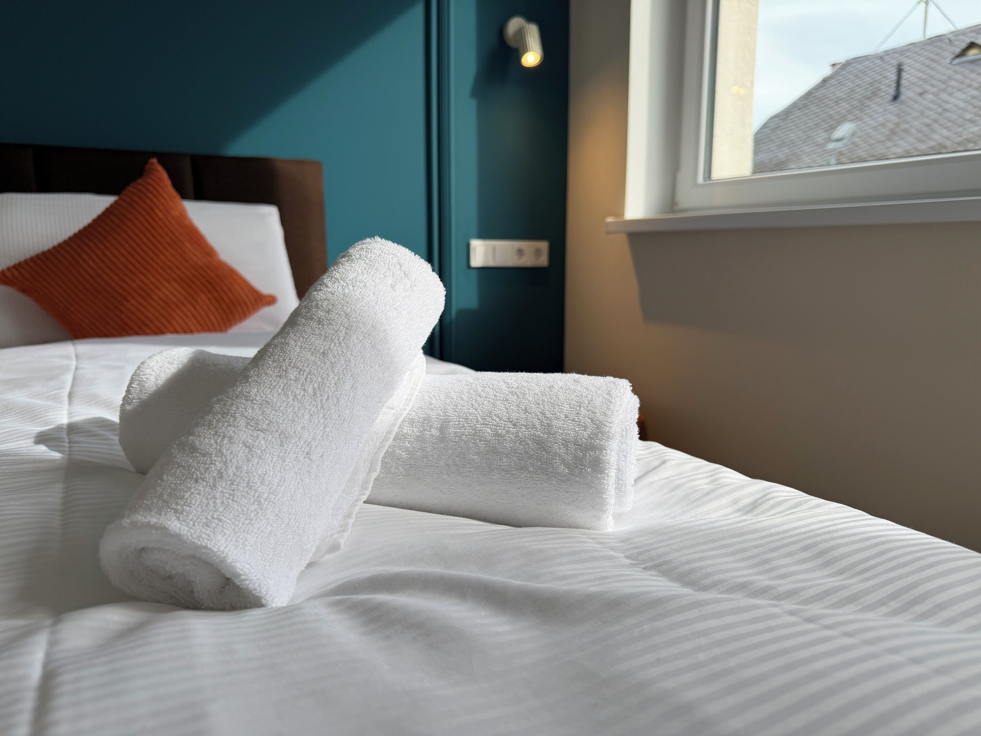 A neatly made bed with white linens and a rolled white towel, a teal-colored wall, and a window with a view of a building.