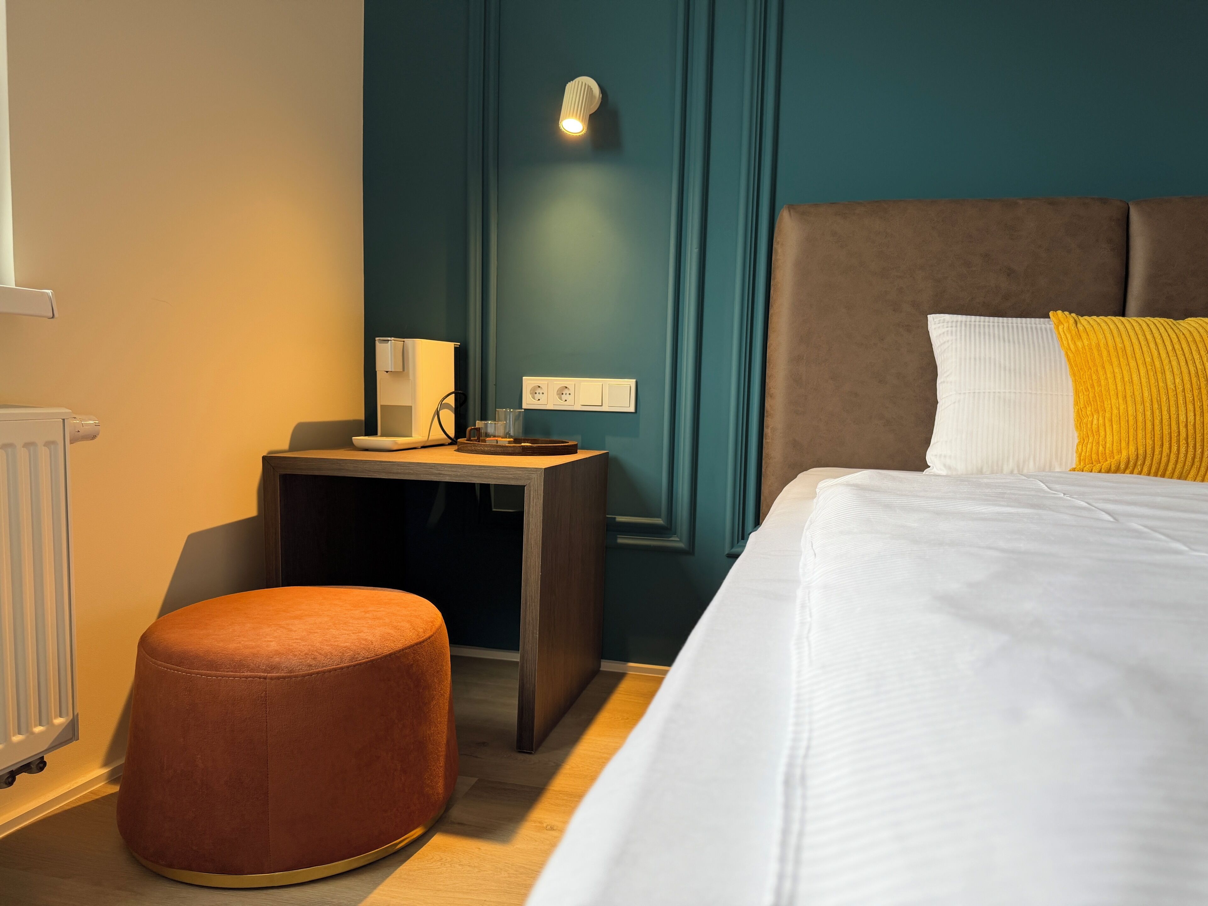 Una habitación de hotel con cama, una mesita con cafetera, un taburete redondo y una lámpara en la pared.