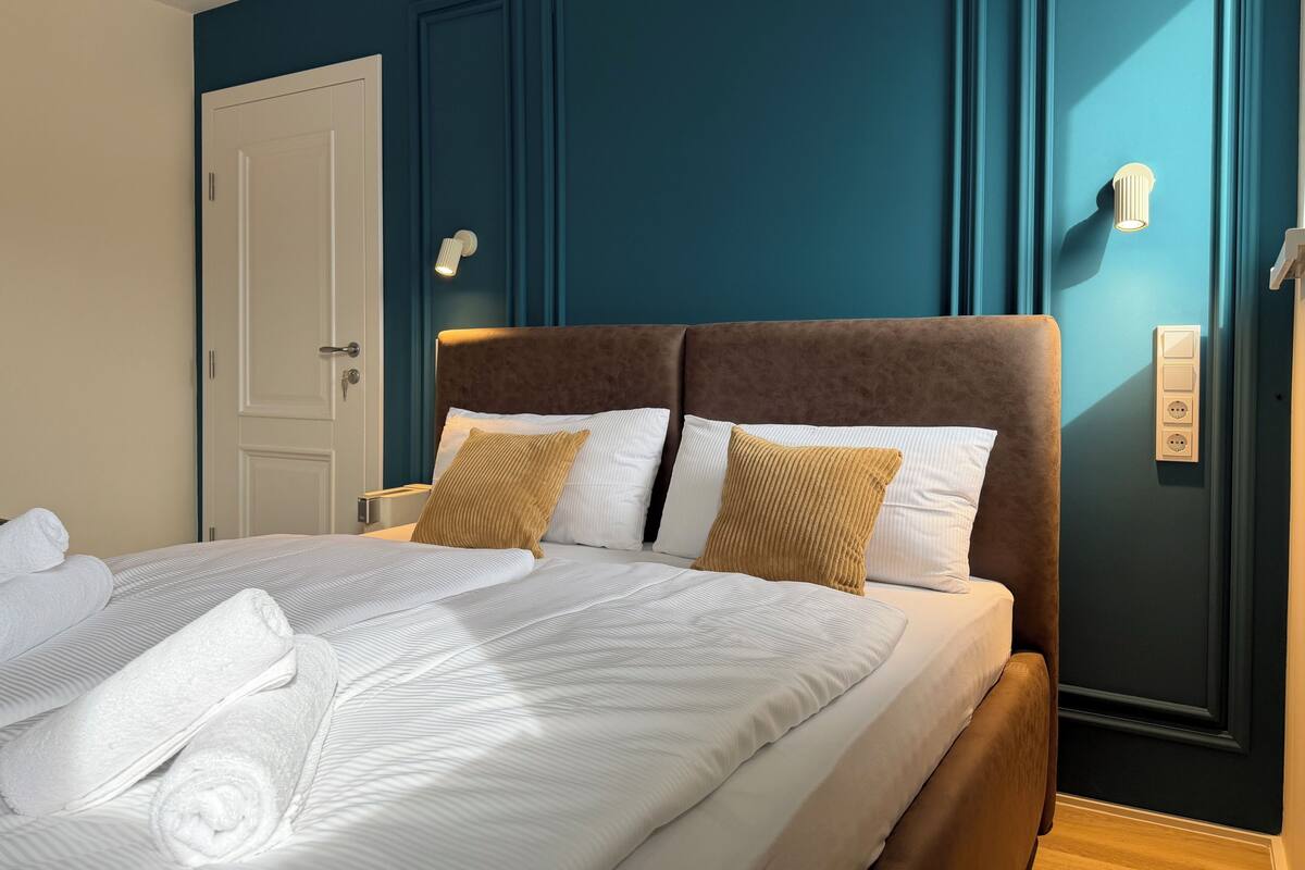 A neatly made bed with white linens and two brown pillows, a bedside table with a lamp, and a door with a handle.