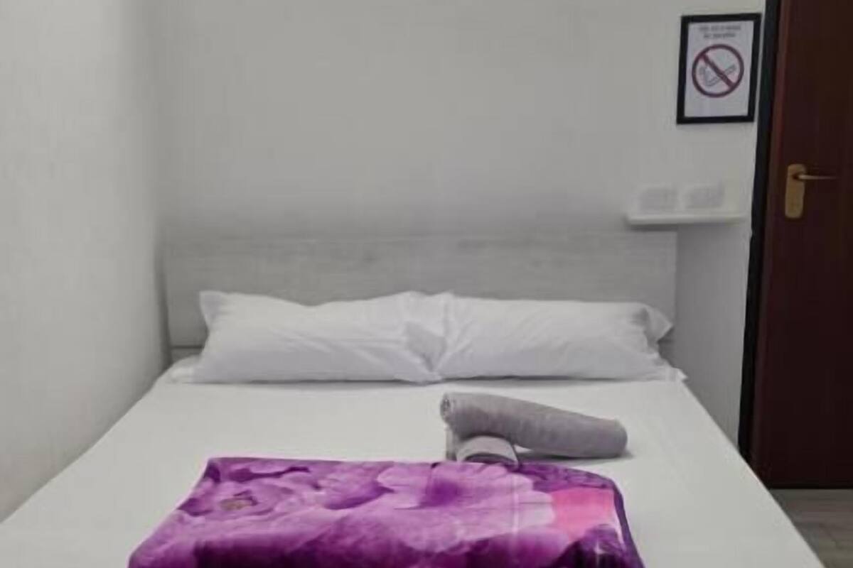 A single bed with white bedding and a purple blanket, a white pillow, and a small bedside table with a lamp. There is a door to the right and a framed sign on the wall.